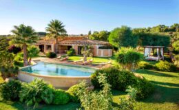 Luxurious villa with a swimming pool surrounded by lush greenery, palm trees, and a shaded outdoor seating area, set against a clear blue sky. Ideal for relaxation and outdoor living.