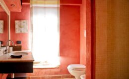 Modern bathroom interior featuring red textured walls, a wooden vanity with a sink, and a white toilet. Natural light streams in through a window with sheer curtains, enhancing the cozy atmosphere.