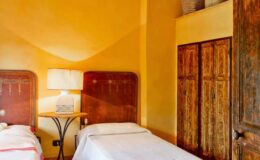 Cozy bedroom with two twin beds, warm yellow walls, a stylish lamp on a side table, and a wooden wardrobe, creating a welcoming and relaxed atmosphere.