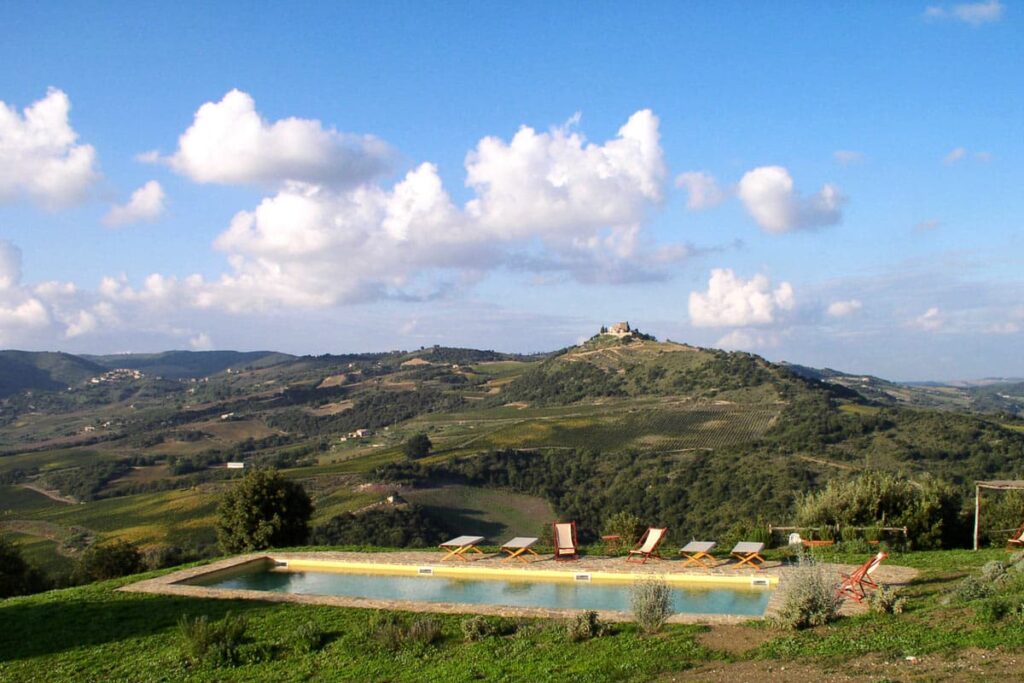 Scenic view of a countryside landscape featuring a swimming pool in the foreground, surrounded by rolling hills and a distant castle on a hilltop under a blue sky with fluffy clouds. | Only on SopranoVillas
