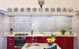 Bright and modern kitchen featuring red cabinets, a marble countertop, and decorative ceramic plates on the wall. The blue-tiled backsplash adds a pop of color, while a lantern light fixture enhances the inviting atmosphere. Fresh fruit is displayed on the counter, showcasing a blend of style and functionality.