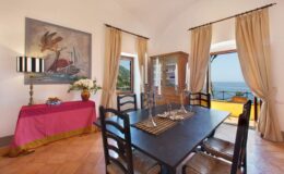 Cozy dining area with a wooden table, six chairs, and elegant decor, featuring a vibrant painting, a sideboard, and large windows offering a view of the sea. Natural light floods the room through sheer curtains.