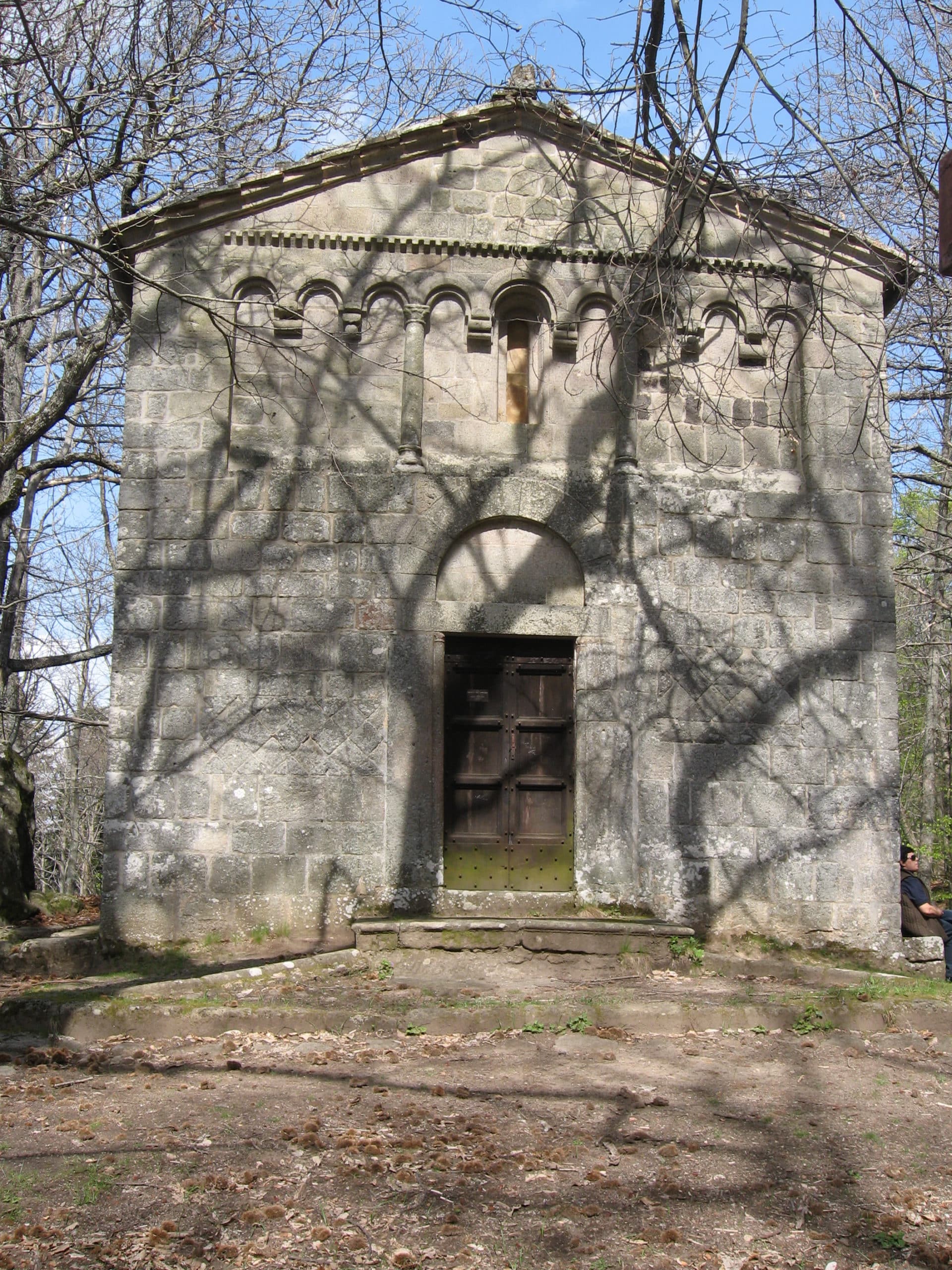 Abandoned stone building with a large wooden door, surrounded by trees casting intricate shadows on the facade. Ideal for exploring historical architecture and nature.