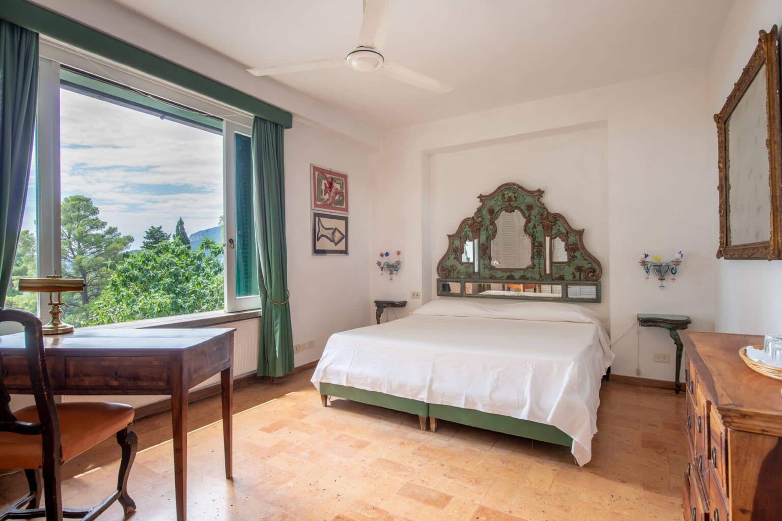 Cozy bedroom with a large window showcasing scenic views, featuring a double bed with an ornate headboard, a wooden desk, and vintage decor elements. | Only on SopranoVillas