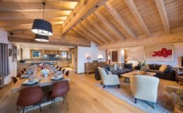 Spacious modern living room and dining area with wooden beams, stylish furniture, and a warm color palette, featuring a large dining table set for a meal and a cozy seating arrangement.