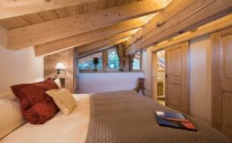 Cozy bedroom with wooden beams, featuring a comfortable bed adorned with decorative pillows, a bedside lamp, and a view of nature through a large window. The room includes rustic wooden doors and warm lighting, creating a welcoming atmosphere.