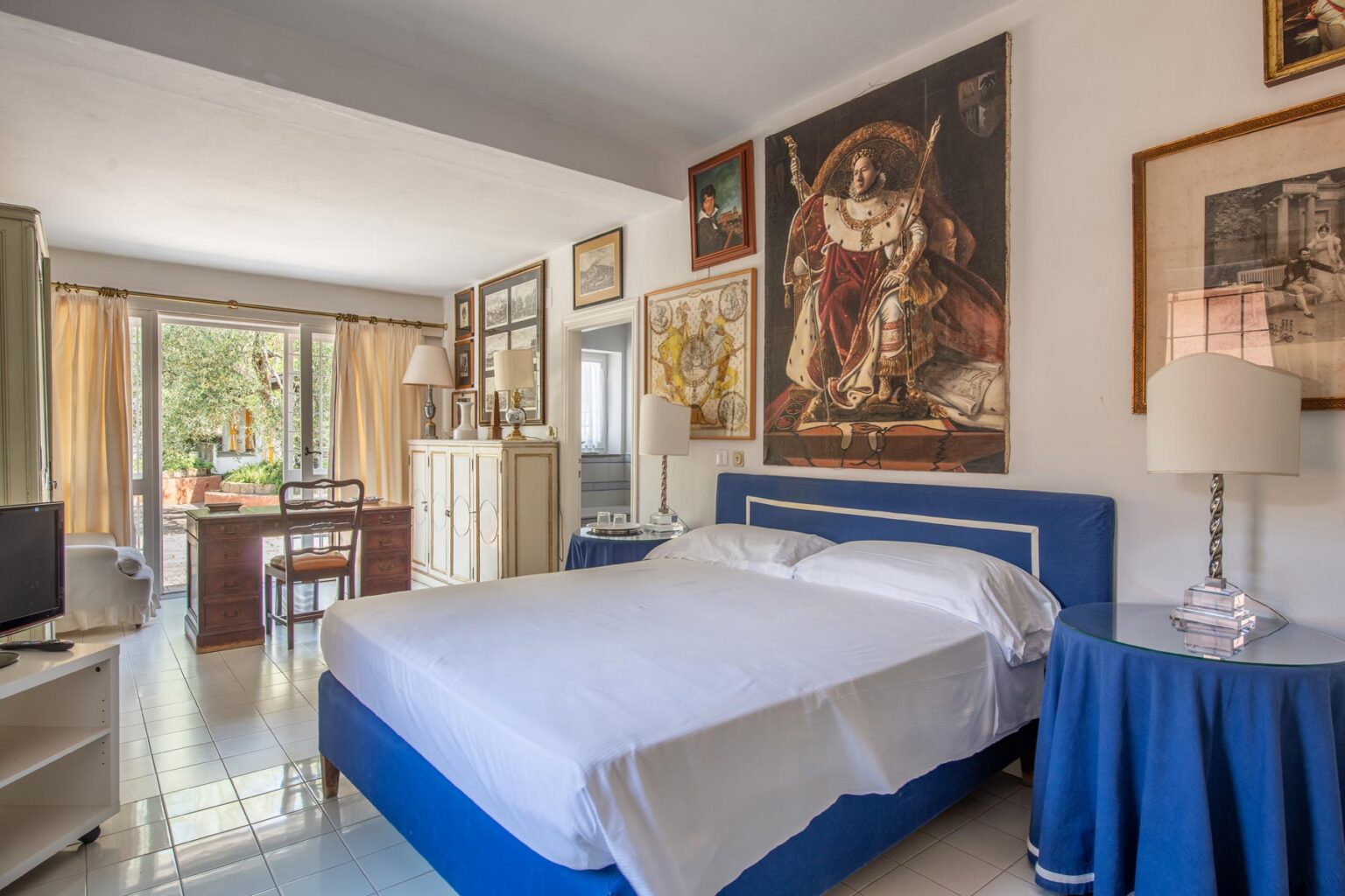 Cozy bedroom featuring a blue bed with white linens, stylish lamps, and a large artwork on the wall. The room is well-lit with natural light from large windows, complemented by elegant decor and a small workspace. | Only on SopranoVillas