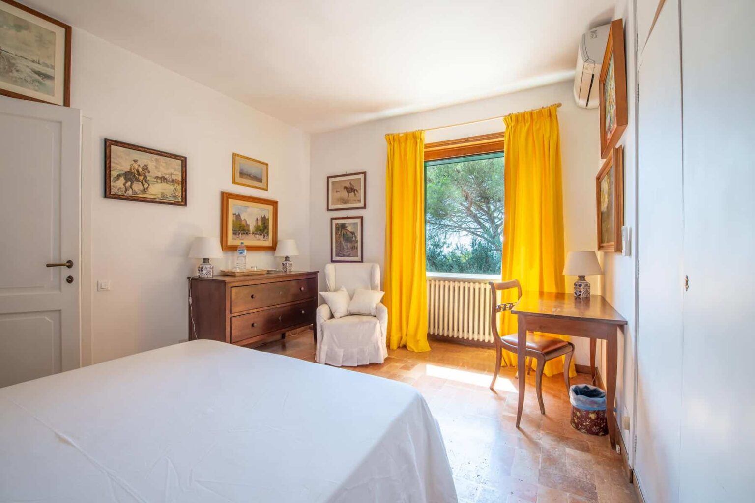 Bright and cozy bedroom featuring yellow curtains, a wooden desk, and vintage decor. Sunlight streams through the window, illuminating the space and highlighting the simple, inviting furnishings. | Only on SopranoVillas