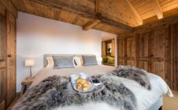 Cozy bedroom featuring a king-sized bed with plush bedding, rustic wooden beams, and warm lighting. A tray with croissants and coffee sits on the bed, enhancing the inviting atmosphere.