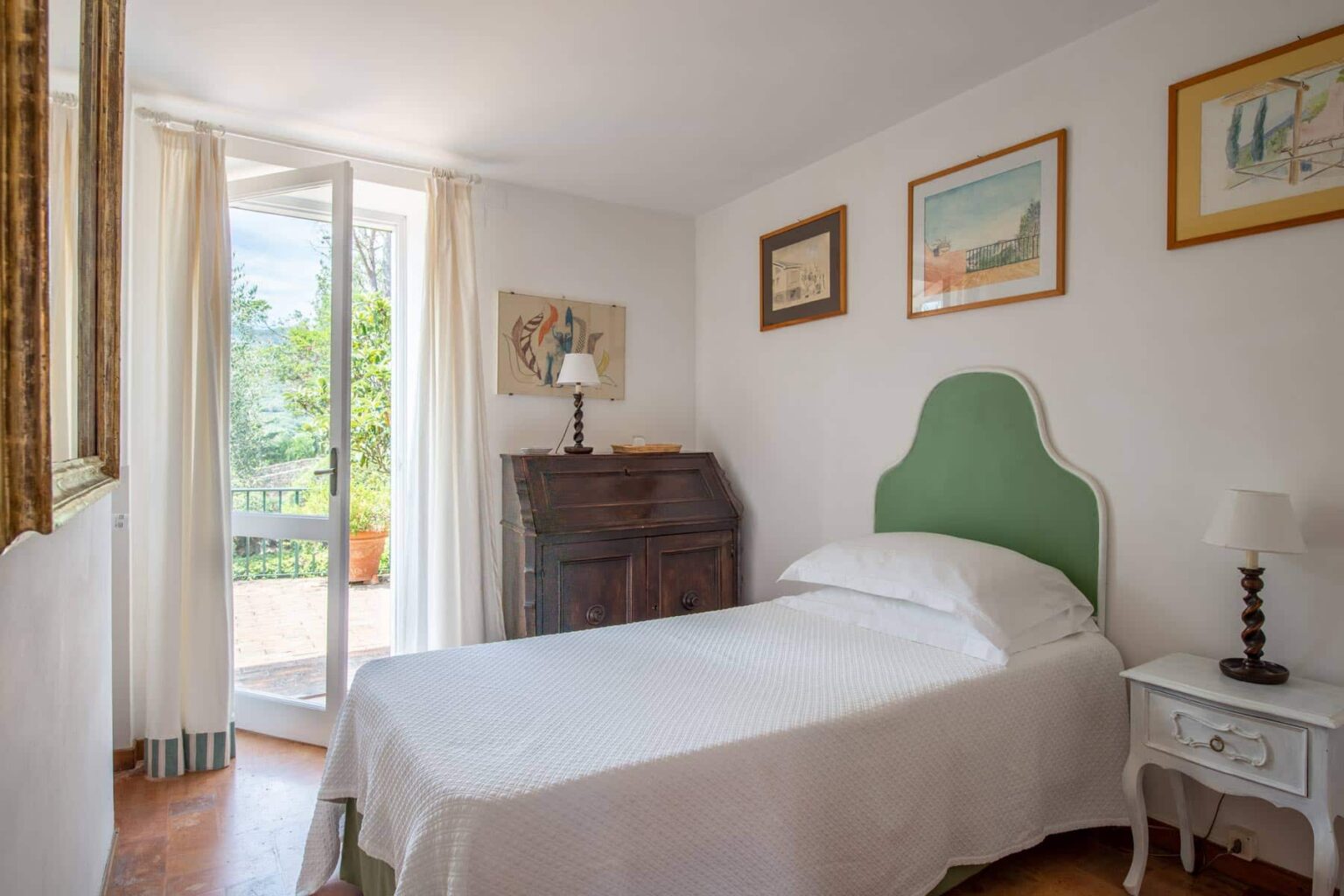 Cozy bedroom featuring a single bed with a white bedspread, green headboard, and light-colored walls. The room includes a vintage wooden desk, a bedside table with a lamp, and artwork on the walls. Large glass doors open to a bright outdoor view, enhancing the airy ambiance. | Only on SopranoVillas