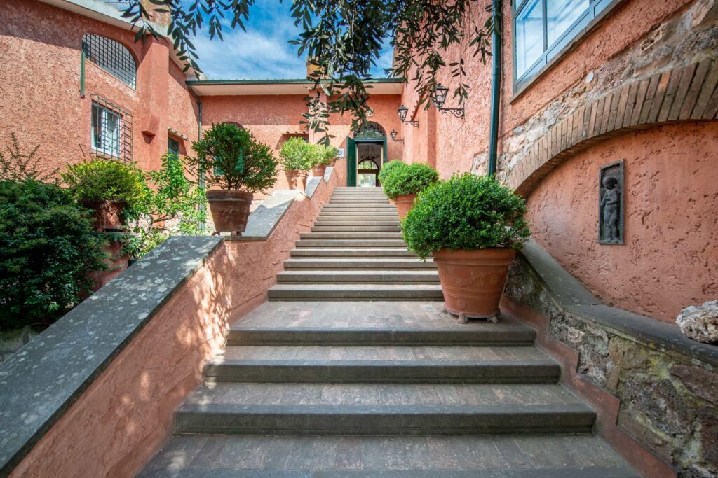Stone steps leading to a charming entrance of a pink building, flanked by potted shrubs and greenery, under a clear blue sky. | Only on SopranoVillas