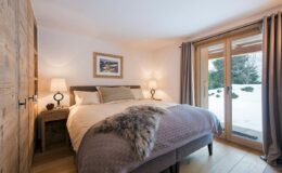 Cozy bedroom featuring a comfortable queen-sized bed with soft bedding, wooden nightstands with lamps, and large windows offering a view of a snowy landscape. The room has warm lighting and rustic decor elements, creating a serene atmosphere.
