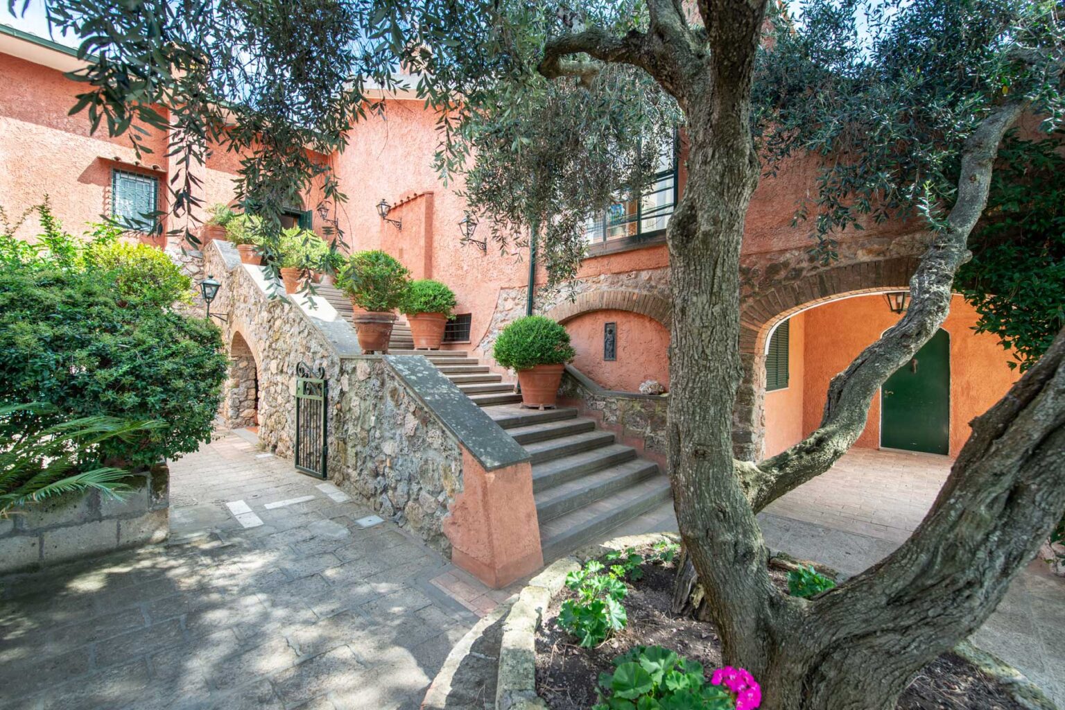Charming outdoor courtyard featuring stone steps, vibrant potted plants, and olive trees, set against a backdrop of a rustic orange building. Ideal for showcasing Mediterranean architecture and serene garden spaces. | Only on SopranoVillas