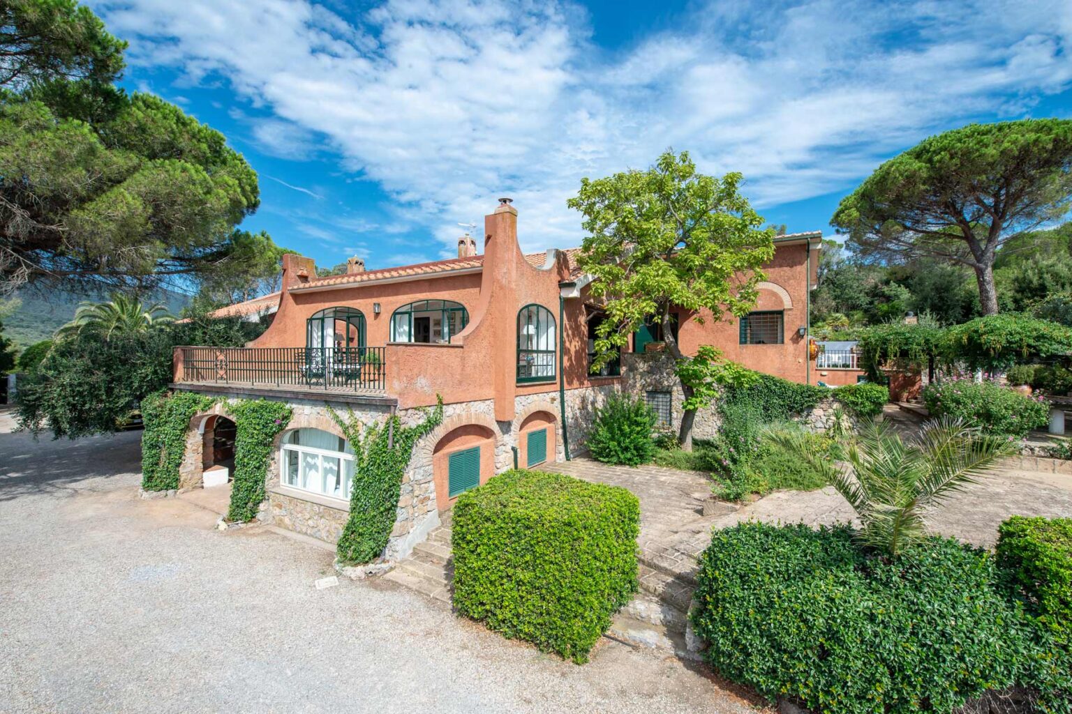 Charming Mediterranean villa surrounded by lush greenery and trees, featuring a unique architectural design and a spacious outdoor area, set against a clear blue sky. | Only on SopranoVillas
