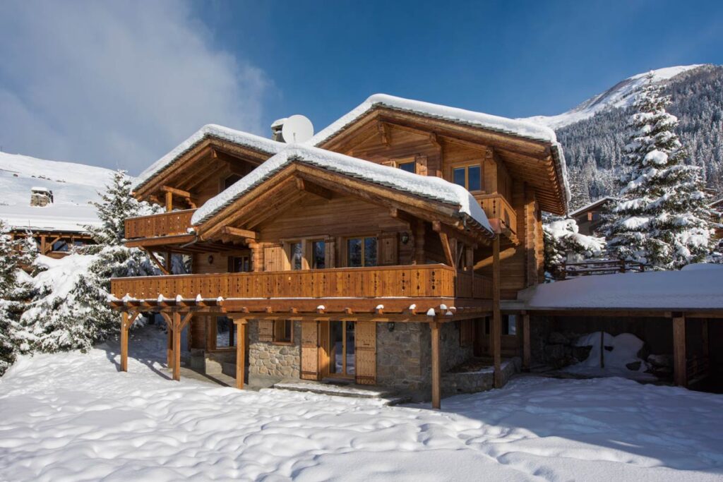 Charming wooden chalet in a snowy landscape, featuring a spacious balcony and surrounded by evergreen trees, set against a backdrop of mountains under a clear blue sky. Ideal for winter retreats and holiday getaways. | Only on SopranoVillas