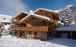 Charming wooden chalet in a snowy landscape, featuring a spacious balcony and surrounded by evergreen trees, set against a backdrop of mountains under a clear blue sky. Ideal for winter retreats and holiday getaways.
