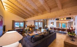 Modern living room interior with wooden beams, plush seating, and a cozy dining area. Natural light floods the space through large windows, creating an inviting atmosphere. Decor includes stylish furnishings and warm accents, ideal for relaxation and entertaining.