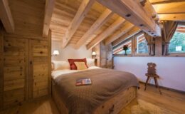 Cozy wooden bedroom with sloped ceiling, featuring a double bed with warm bedding, bedside lamps, and a small reading chair, surrounded by natural light from large windows.
