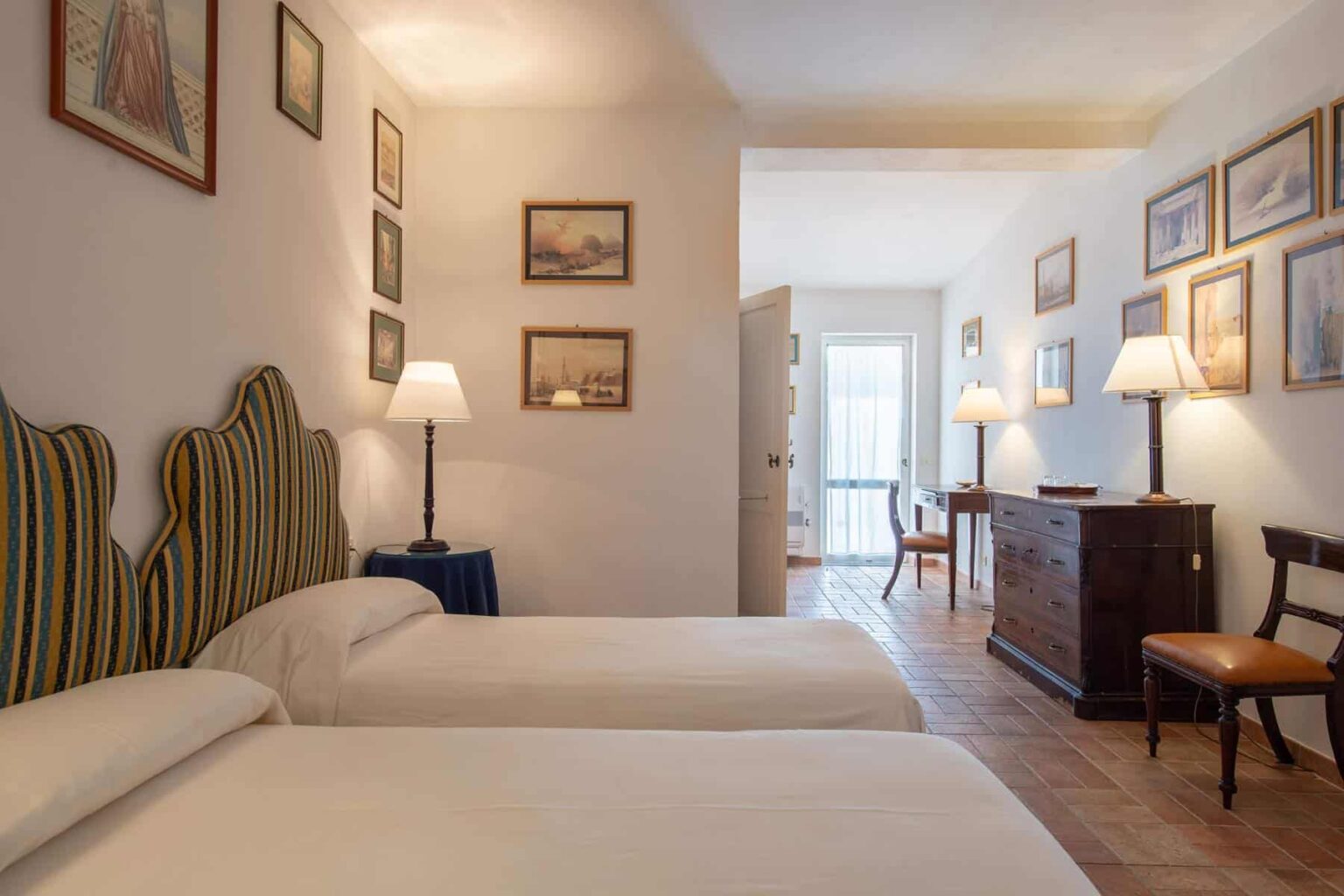 Cozy hotel room featuring two twin beds with striped headboards, warm lighting from bedside lamps, and art-adorned walls, creating a welcoming atmosphere for guests. | Only on SopranoVillas