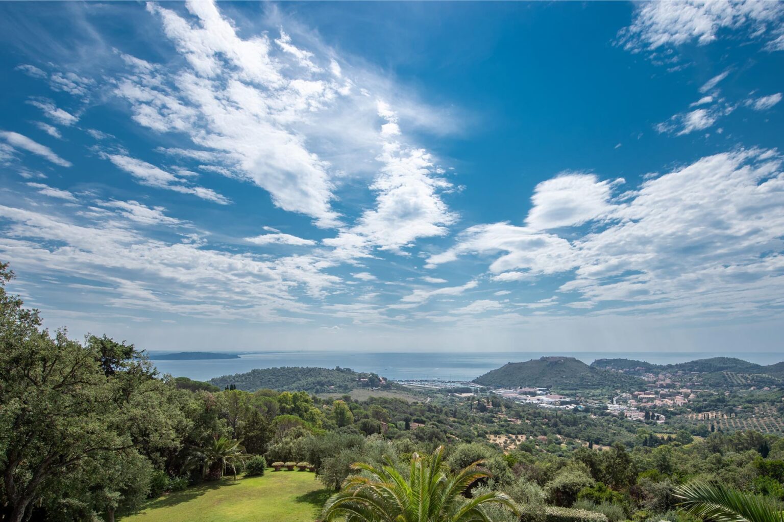 Scenic view of a coastal landscape featuring rolling hills, lush greenery, and a vibrant blue sky with scattered clouds, overlooking the sea and a nearby town. | Only on SopranoVillas