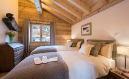 Cozy bedroom with two twin beds, wooden accents, and warm lighting in a chalet-style interior, featuring a dresser, decorative artwork, and soft bedding. Ideal for a relaxing getaway.