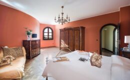 Elegant bedroom featuring a white bed with decorative pillows, a vintage wooden wardrobe, a cozy seating area, and warm orange walls, illuminated by a stylish chandelier.