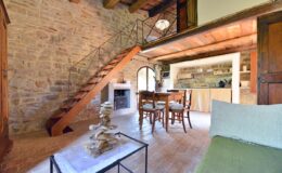 Cozy rustic living area featuring a stone wall, wooden staircase, dining table with chairs, and a modern kitchen. Natural light illuminates the space, highlighting the blend of traditional and contemporary design elements.