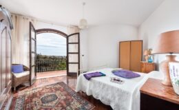 Cozy bedroom with a double bed, wooden furniture, and large windows offering a scenic view. The room features a colorful rug and decorative pillows, creating a warm and inviting atmosphere.