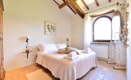 Cozy bedroom with wooden beams, a large window, and a comfortable bed with white linens and decorative pillows, featuring minimalist decor and warm natural light.
