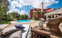 Luxury outdoor living space featuring a swimming pool, comfortable lounge chairs, and a cozy patio area surrounded by lush greenery and vibrant landscaping. The scene is set against a backdrop of a stylish home under a bright blue sky with fluffy clouds.