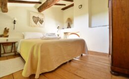 Cozy bedroom featuring a comfortable bed with a beige blanket, wooden beams on the ceiling, and warm wooden flooring, creating a welcoming ambiance. Decor includes wall art and soft lighting, enhancing the serene atmosphere.