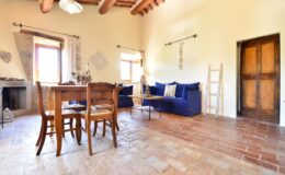 Cozy living room featuring a wooden dining table with chairs, a blue sofa, and rustic decor. The room has terracotta flooring, wooden beams, and large windows that allow natural light to fill the space.