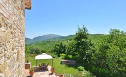 Scenic view from a stone villa overlooking lush green hills and a clear blue sky, featuring a shaded outdoor seating area with a gazebo.