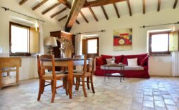 Cozy living room with wooden beams, a red sofa, wooden dining table, and natural light from windows, showcasing rustic decor and a warm atmosphere.