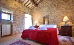 Cozy bedroom with stone walls, a red bedspread, and two white towels on the bed. Features include wooden furniture, a window with natural light, and a warm ambiance. Ideal for a relaxing getaway.