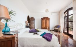 Bright and airy bedroom featuring a comfortable bed with colorful pillows, a wooden wardrobe, a cozy reading chair, and large windows that let in natural light. The decor includes a stylish lamp and indoor plants, creating a relaxing atmosphere.