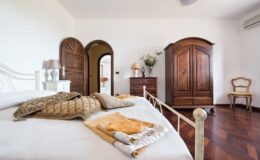 Cozy bedroom interior featuring a white bed with decorative pillows and blankets, a wooden wardrobe, a dresser, and a vintage chair. Natural light illuminates the space, highlighting the warm wooden flooring and elegant decor elements.