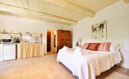 Cozy and inviting bedroom with a white bedspread, wooden ceiling beams, and a small kitchenette, ideal for a relaxing getaway.