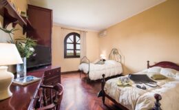 Cozy guest room featuring two twin beds, a wooden desk, and a flat-screen TV, with warm yellow walls and a large window providing natural light. Ideal for travelers seeking comfort and relaxation.