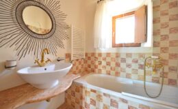 Modern bathroom featuring a stylish bathtub and a contemporary sink with gold fixtures, adorned with a decorative mirror. Natural light enters through a window, highlighting the warm tile design.