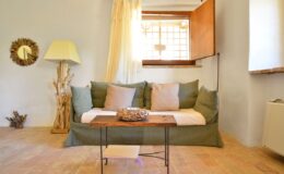 Cozy living room featuring a green sofa with decorative pillows, a wooden coffee table, and a floor lamp, illuminated by natural light from a window with sheer curtains. A mirror and decorative accents enhance the inviting atmosphere.