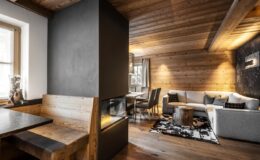 Modern rustic living room featuring a cozy seating area with a grey sofa and wooden accents, alongside a stylish dining nook with a wooden bench. The space is illuminated by warm lighting, highlighting the wooden ceiling and walls for a welcoming atmosphere.