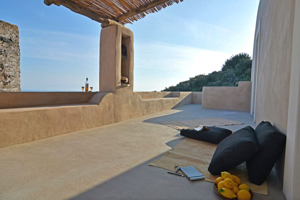 Spacious outdoor terrace with minimalist design, featuring a sandy floor, black cushions, and a glimpse of the ocean in the background, surrounded by greenery and natural stone walls. | Only on SopranoVillas
