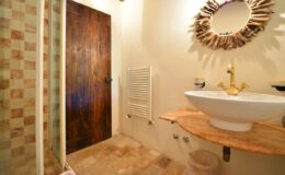 Modern bathroom featuring a wooden door, a stylish round mirror, a sleek white sink on a natural stone countertop, and a decorative radiator. The floor includes patterned tiles, contributing to a warm and inviting atmosphere.