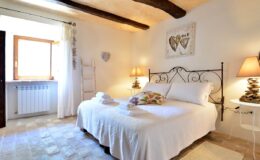Cozy bedroom featuring a double bed with white linens, decorative pillows, and bedside lamps, set in a bright space with wooden beams, a window allowing natural light, and stylish decor elements. Ideal for a relaxing getaway.