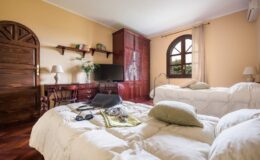 Cozy bedroom featuring two twin beds, a wooden wardrobe, a desk with a chair, and a television. The room has warm yellow walls and large windows, providing natural light and a welcoming atmosphere.