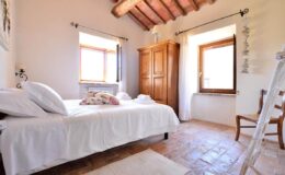 Cozy bedroom with a wooden ceiling, featuring a double bed with white linens, a wooden wardrobe, and a chair. Natural light streams in through the windows, creating a warm and inviting atmosphere.