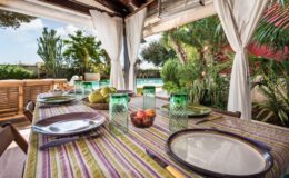 Outdoor dining area with a colorful table setting, featuring plates, glasses, and fresh fruit, surrounded by lush greenery and a view of a swimming pool.