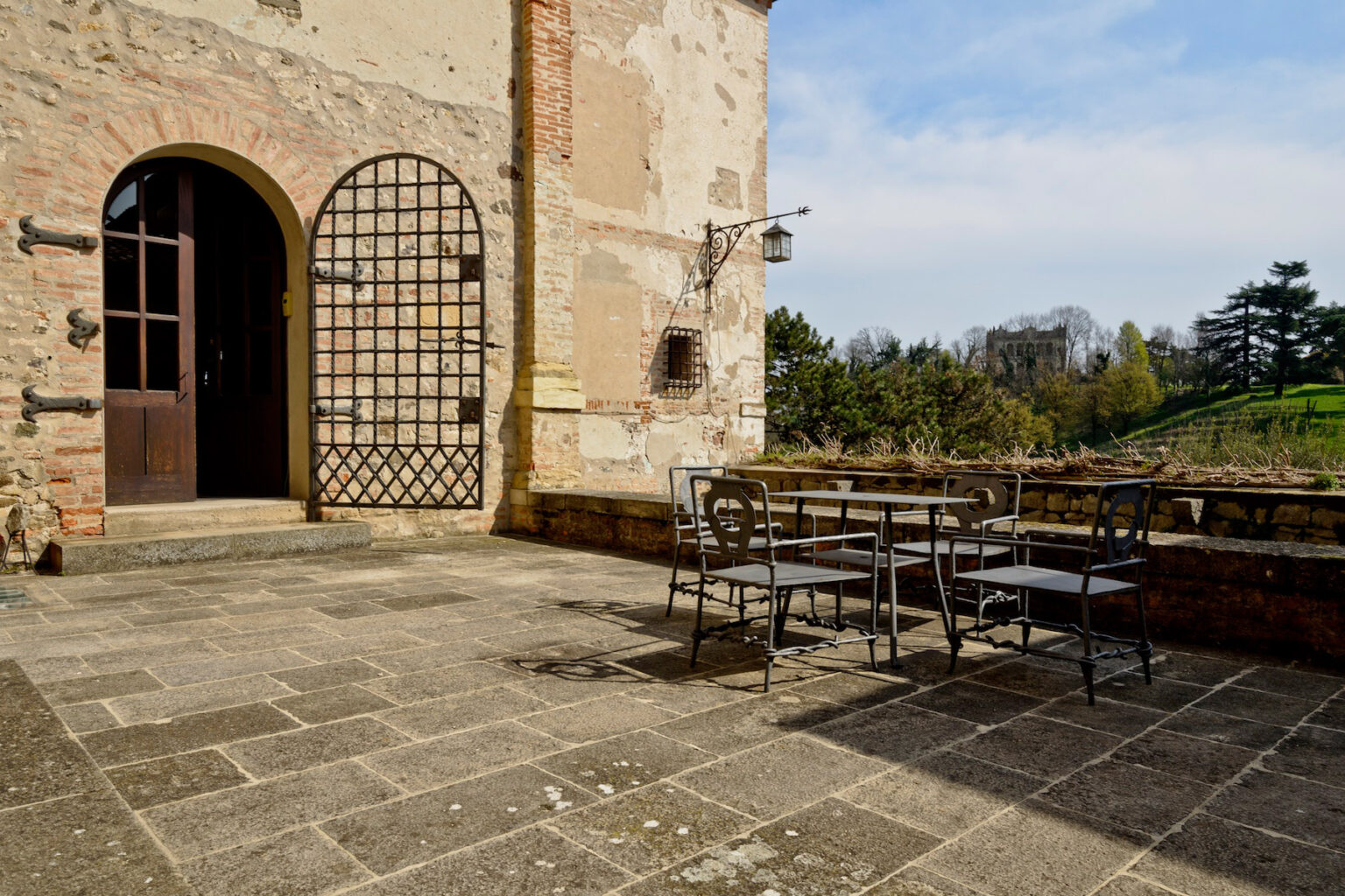 Outdoor patio area featuring a rustic stone floor, a wrought iron table with chairs, and an arched doorway leading to a historic building. Lush greenery and trees are visible in the background under a clear blue sky. | Only on SopranoVillas