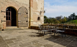 Outdoor patio area featuring a rustic stone floor, a wrought iron table with chairs, and an arched doorway leading to a historic building. Lush greenery and trees are visible in the background under a clear blue sky.